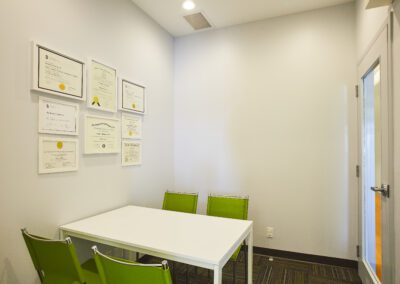 A clean consultation room with certificates on the wall, a white table, and three bright green chairs.